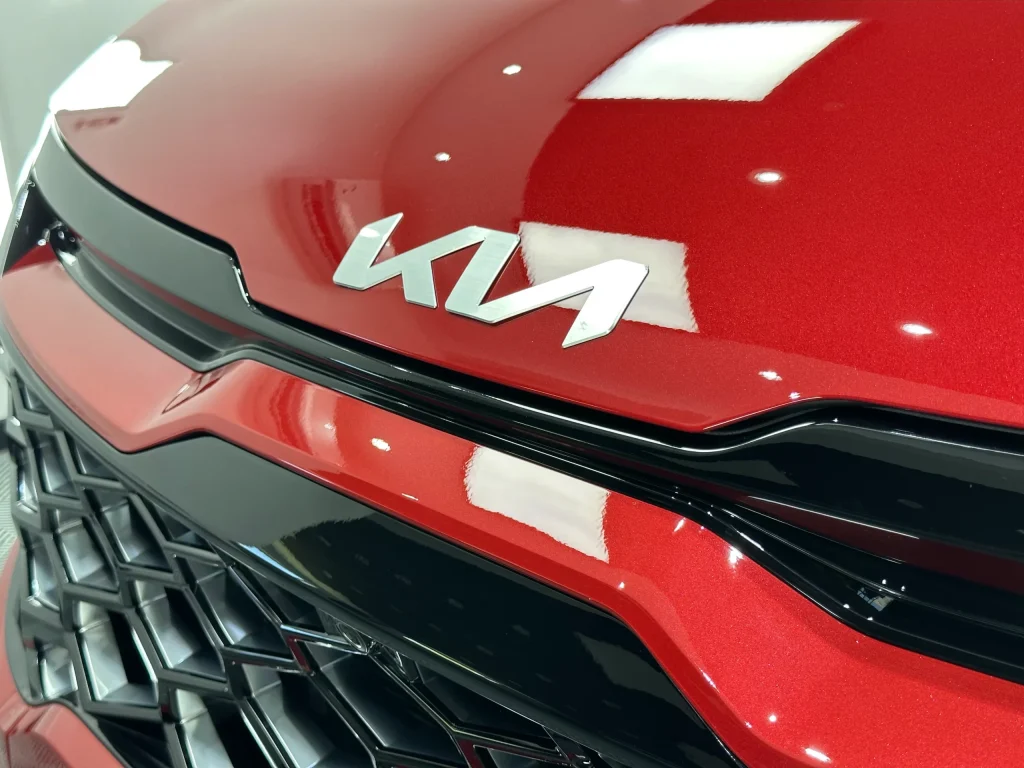 Sigla Kia și marginea capotei la un Kia Sportage roșu, după aplicarea foliei PPF